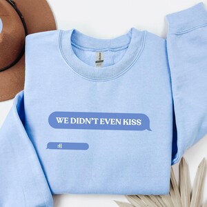 Può includere: Felpa azzurra con la scritta "WE DIDN'T EVEN KISS" in un fumetto e un'emoji triste. La felpa è di colore azzurro chiaro. Un cappello marrone è parzialmente visibile.
