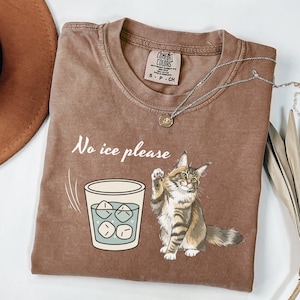 Pode incluir: Camiseta marrom com a frase "No ice please" e um desenho de um gato acenando para um copo com cubos de gelo. A camiseta tem gola redonda e uma etiqueta pequena. Um colar de prata com um pingente também é visível.