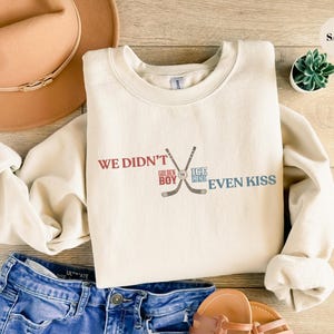 Può includere: Felpa color sabbia con la scritta "WE DIDN'T EVEN KISS" in rosso e blu, con bastoni da hockey incrociati. La felpa è appoggiata su una superficie di legno con jeans blu, un cappello marrone e sandali.