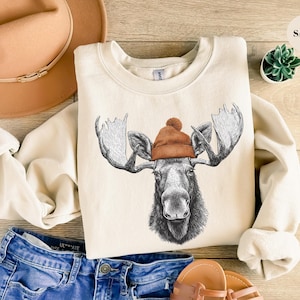 Könnte beinhalten: Cremefarbenes Sweatshirt mit einer detaillierten Illustration eines Elchs mit einer braunen Strickmütze. Der Elch hat ein großes Geweih und ein detailliertes Gesicht. Das Sweatshirt wird mit blauen Jeansshorts und einem beigen Hut kombiniert.