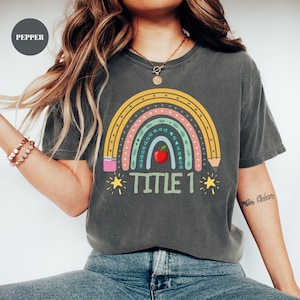 May include: Dark gray t-shirt featuring a colorful rainbow design with a red apple at the center. The text "TITLE 1" is printed below the rainbow, flanked by yellow stars. The shirt is a casual style.