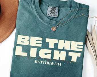 May include: A teal Comfort Colors t-shirt with the words "BE THE LIGHT" in large, off-white block letters. Below the text is "MATTHEW 5:14" and a string of colorful lights. A gold necklace with a pendant rests on the shirt.