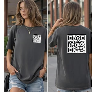 Könnte beinhalten: Anthrazitfarbenes T-Shirt mit einem weißen QR-Code auf der Vorder- und Rückseite. Das Shirt hat kurze Ärmel und eine lockere Passform. Das Model trägt Jeansshorts und eine Halskette.