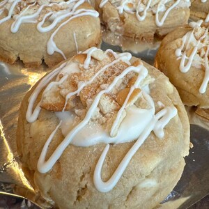 May include: Close-up of several baked cookies with a light brown exterior and white icing drizzle. The cookies have a creamy white filling and are topped with crumbled pieces. The cookies are arranged on a silver surface.