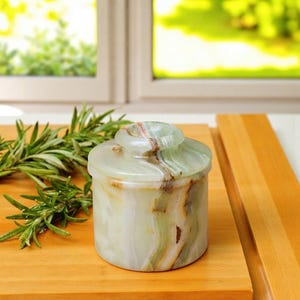 Pode incluir: Um recipiente cilíndrico de pedra ónix verde claro com tampa a condizer, apresentando padrões naturais de veios minerais brancos, creme e castanhos em toda a superfície polida, servindo como um recipiente de armazenamento decorativo.