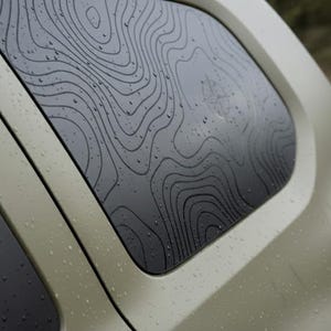 Può includere: Primo piano del finestrino posteriore di un veicolo con un design a mappa topografica. Il finestrino è scuro con un motivo a contorno nero e la carrozzeria dell'auto è verde tenue. Sono visibili goccioline d'acqua.