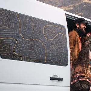 Puede incluir: Furgoneta blanca con una ventana oscura con un diseño de mapa topográfico dorado. La puerta de la furgoneta está abierta, revelando personas en el interior. El fondo muestra un paisaje montañoso.