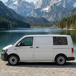 Op de afbeelding: Een witte bestelwagen geparkeerd bij een meer met bergen op de achtergrond. De bestelwagen heeft een zwart-wit topografisch kaartontwerp op de achterruit. De bestelwagen staat op een grindoppervlak.