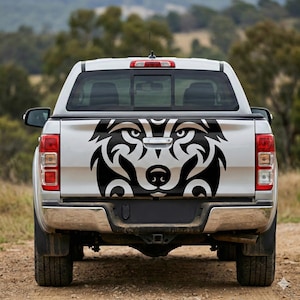 Puede incluir: Una camioneta blanca con una calcomanía de cabeza de lobo tribal negra en el portón trasero. El diseño del lobo presenta patrones y detalles intrincados, con las luces traseras y el parachoques de la camioneta visibles.