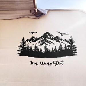 Op de afbeelding: Zwarte berg- en bossticker met twee vogels die eroverheen vliegen. De sticker is aangebracht op de zijkant van een camper. De tekst "Dein Wunschtext" staat eronder in cursief.