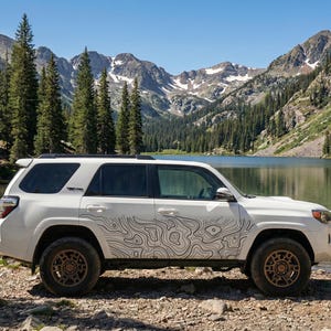 Può includere: SUV bianco con un design a mappa topografica sul lato, parcheggiato vicino a un lago. Il veicolo ha ruote color bronzo ed è ambientato sullo sfondo di montagne e un cielo azzurro. La scena evoca avventura ed esplorazione all'aperto.