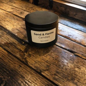 May include: A black candle tin with a lid, labeled "Sand & Farms Candles" with "Beach Heather" below. The candle sits on a rustic wooden surface, with a window in the background.