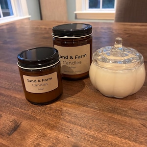 Può includere: Due barattoli in vetro ambrato con coperchi neri ed etichette beige con la scritta Sand & Farm Candles Foliage Hand Poured Soy Wax, accanto a un contenitore in vetro trasparente a forma di zucca riempito di cera bianca con coperchio.