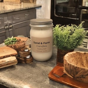 Op de afbeelding: Een potkaars met een zilveren deksel en een etiket met de tekst "Sand & Farm Candles Strawberry Patch". De kaars is omgeven door houten kommen en een kleine plant. De kaars is met de hand gegoten en gemaakt van sojawas.