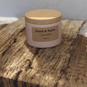 Può includere: Una candela cilindrica in un contenitore rosa chiaro con un coperchio in legno chiaro. Un'etichetta rettangolare color cuoio sul davanti presenta il testo nero Sand & Farm Candles e la parola Driftwood stampata in piccolo sotto il marchio.