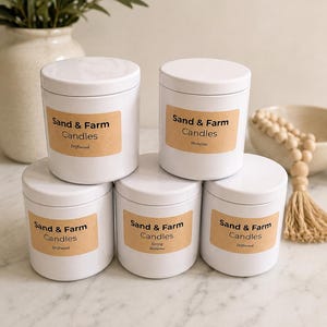 Può includere: Cinque barattoli cilindrici bianchi per candele con coperchi abbinati, ognuno dotato di un'etichetta rettangolare color marrone chiaro con il testo Sand & Farm Candles e nomi di fragranze come Driftwood, Shoreline e Spring Blossoms.