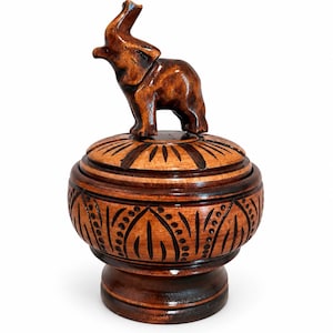 Puede incluir: Un recipiente redondo de madera de color marrón oscuro con tapa, que presenta una figura de elefante tallada en la parte superior. La tapa y el cuerpo del recipiente están decorados con patrones oscuros grabados de líneas y puntos.