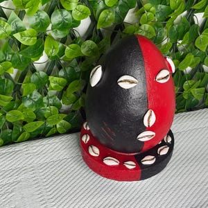 Pode incluir: Uma escultura em forma de ovo dividida em metades preta e vermelha, decorada com conchas de búzios brancas. As conchas estão dispostas na superfície do ovo e ao redor da base circular, também dividida em seções preta e vermelha correspondentes.