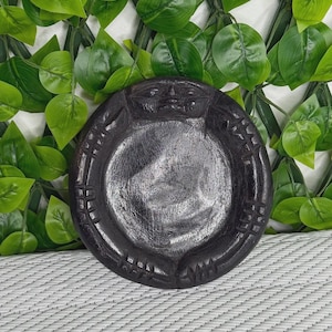 Puede incluir: Un cuenco circular de madera de color marrón oscuro con un rostro tallado en el borde superior y marcas grabadas decorativas a lo largo del borde exterior. El cuenco tiene una superficie interior lisa y pulida y una base profunda y redondeada.