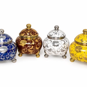Puede incluir: Cuatro tarros redondeados con tapa y tres patas, con motivos florales. Los tarros son azul con plata, marrón con oro, blanco con plata y amarillo con oro. Cada tarro tiene una tapa a juego con un pequeño pomo como asa en la parte superior.
