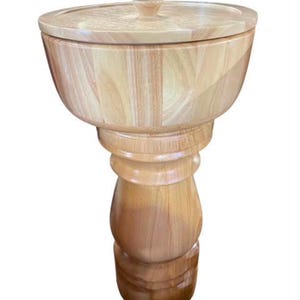 May include: A light-colored wooden pedestal bowl with a matching circular lid featuring a small central knob. The bowl has a wide, rounded upper section that tapers down into a thick, turned support column with a stable, circular base.