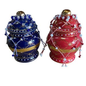 May include: Two decorative ceramic jars, one navy blue and one red, with gold accents and beaded embellishments. Each jar has a gold-colored knob on top and a beaded strand draped around the lid. The jars have a starburst pattern.