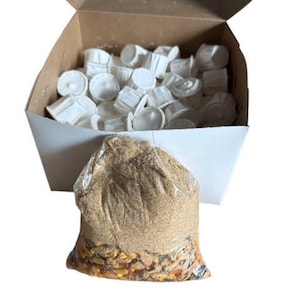 May include: An open cardboard box containing numerous white, cylindrical items. In front of the box is a clear plastic bag filled with tan and brown granules and seeds. The box and bag are set against a white background.