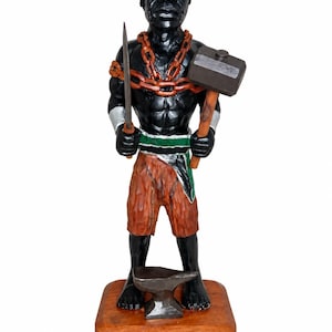 Puede incluir: Una figura de madera tallada a mano de una persona de piel negra, con una gorra verde y negra, pantalones cortos naranjas y una cadena al cuello. La figura sostiene una herramienta de metal y un martillo, sobre una base de madera con un yunque.