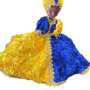 May include: A decorative doll in a vibrant yellow and blue dress, adorned with gold trim. The doll's head is covered with a matching yellow head wrap, embellished with blue jewels. The dress features a rose-like texture.