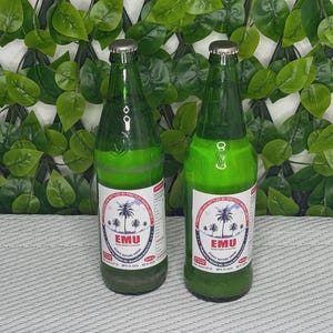 Puede incluir: Dos botellas de vidrio verde de vino de palma Emu con tapas plateadas. Cada botella presenta una etiqueta circular blanca con una ilustración de palmeras, el texto Emu y la instrucción de agitar antes de beber.