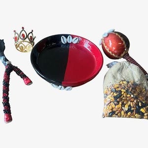 May include: An assortment of spiritual items, including a black and red bowl with cowrie shells, a red and gold crown, a beaded wand, a red maraca, and a bag of grains. The items are arranged on a white background.