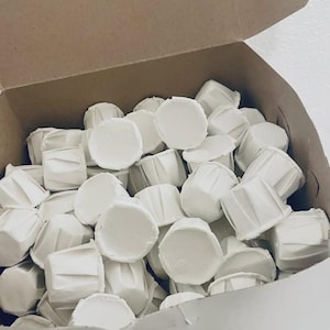 May include: A cardboard box filled with numerous small, white, pleated paper baking cups. The cups are arranged in a jumbled pile, each featuring a circular base and vertically ribbed sides, designed for holding individual portions of baked goods.