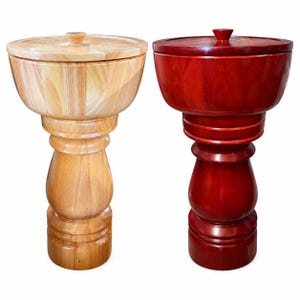 May include: Two tall wooden pepper mills with a smooth, polished finish. One mill features a light, natural wood grain, while the other is stained a deep, rich red. Both have a wide, bowl-shaped top with a lid and a sturdy, pedestal-style base.