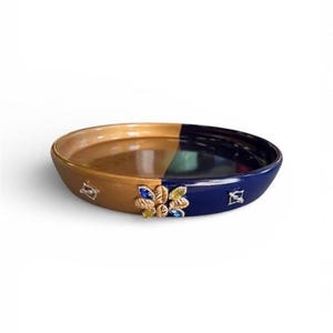 May include: A circular tray featuring a two-tone design with one half in metallic gold and the other in deep navy blue. The exterior rim is decorated with a central cluster of gold-toned leaf shapes accented by blue and yellow crystals.