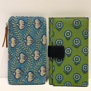 May include: Two fabric wallets. One is teal with a repeating pattern of stylized fish and leaves. The other is green with a pattern of blue and white circles. Both have a rectangular shape. The teal wallet has a zip and a brown leather pull.