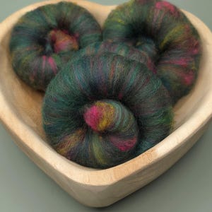 May include: Three coiled bundles of multi-colored wool roving featuring deep forest green, vibrant magenta, and golden yellow fibers. The wool is arranged inside a smooth, light-toned wooden bowl shaped like a heart, showcasing a soft texture.
