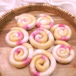 May include: A wooden tray holds eight swirled fiber art pieces. Each spiral features a blend of white, yellow, and pink hues. The tray has a beaded edge and sits on a light-colored surface.
