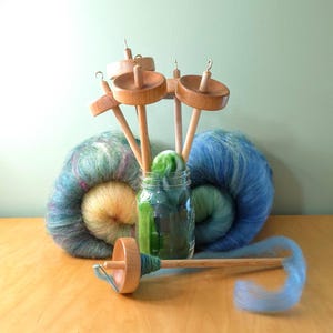 May include: Several wooden drop spindles with metal hooks are arranged in a clear glass jar. Two large, round balls of multi-colored wool roving in shades of blue, green, and yellow sit behind the jar, with one spindle lying horizontally in front.