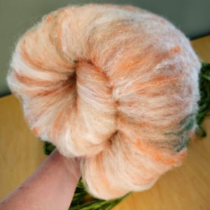 May include: A large, round ball of peach-colored wool roving with streaks of white, orange, and green. The wool is soft and fluffy, ideal for fiber art projects like needle felting or spinning. A hand is holding the wool.