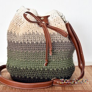 May include: A handmade crochet bucket bag in a gradient of colors, from cream to olive green. It features brown leather accents, including a drawstring closure and a shoulder strap. The bag is designed with a textured stitch.