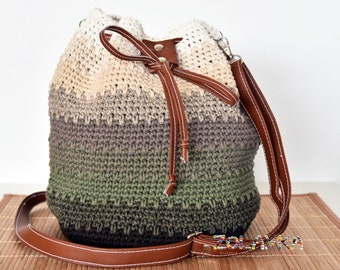 May include: A handmade crochet bucket bag in a gradient of colours, from cream to olive green. It features brown leather accents, including a drawstring closure and a shoulder strap. The bag is designed with a textured stitch.