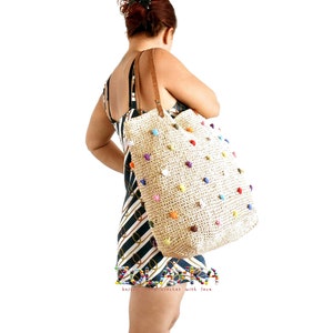 May include: A woman in a black and white striped dress is carrying a large straw tote bag with colorful pom poms.