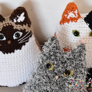 Three crocheted cat toys. One cat is white with brown and tan accents, one is white with orange and black accents, and one is gray with white accents. The gray cat is fuzzy and the other two cats are smooth. The toys are all handmade and have button eyes.
