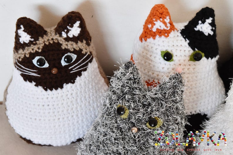 Three crocheted cat toys. One cat is white with brown and tan accents, one is white with orange and black accents, and one is gray with white accents. The gray cat is fuzzy and the other two cats are smooth. The toys are all handmade and have button eyes.