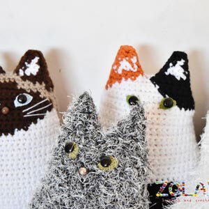 Four crocheted cat toys. One cat is white with brown and beige accents, one is white with black and orange accents, one is white with black accents, and one is gray with white accents. The toys are all sitting on a white surface. The text 'ZOLAYKA' is visible in the bottom right corner of the image. The text 'loving, and crochet with love' is visible below the text 'ZOLAYKA'.