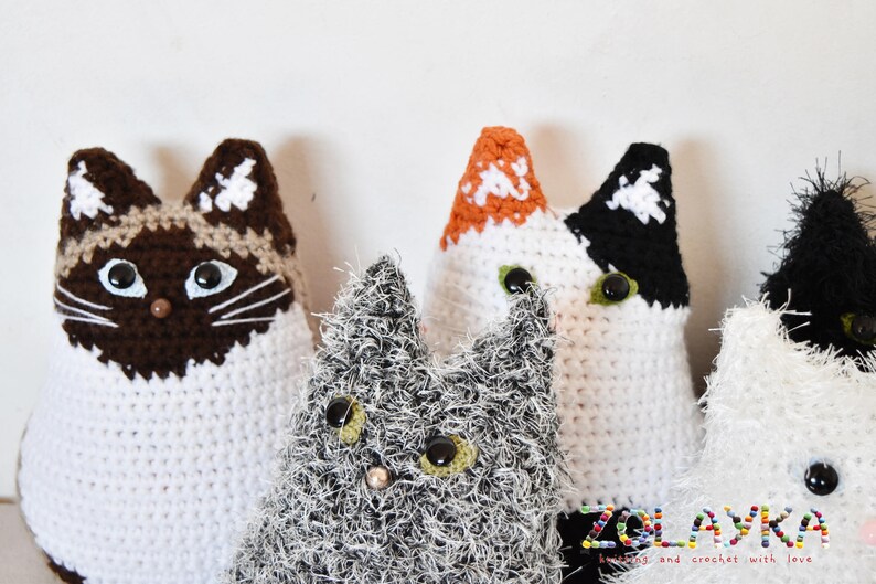 Four crocheted cat toys. One cat is white with brown and beige accents, one is white with black and orange accents, one is white with black accents, and one is gray with white accents. The toys are all sitting on a white surface. The text 'ZOLAYKA' is visible in the bottom right corner of the image. The text 'loving, and crochet with love' is visible below the text 'ZOLAYKA'.