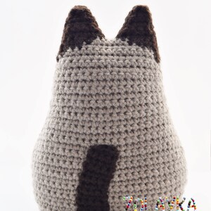 A crocheted stuffed animal in the shape of a cat. The cat is beige with brown ears and a brown tail. The cat is sitting on a white surface. The text 'ZOLASKA knitting & crochet with love' is visible at the bottom of the image.