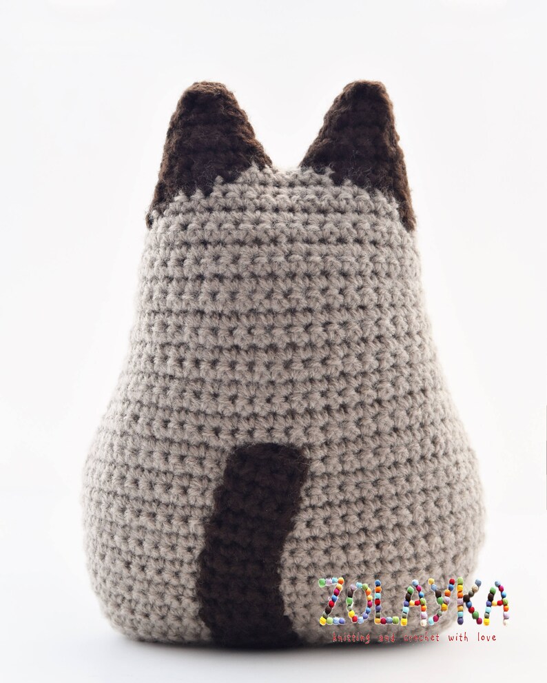 A crocheted stuffed animal in the shape of a cat. The cat is beige with brown ears and a brown tail. The cat is sitting on a white surface. The text 'ZOLASKA knitting & crochet with love' is visible at the bottom of the image.