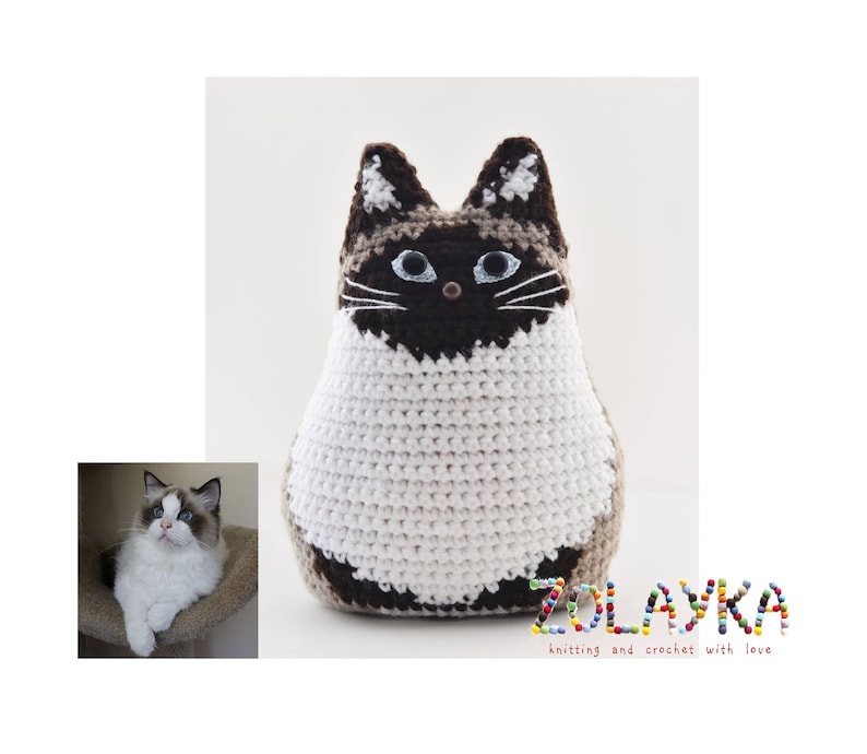 A crocheted stuffed cat toy with brown and white fur. The cat has blue eyes and a black nose. The toy is sitting on a white surface. The text 'ZOLAYKA knitting and crochet with love' is below the toy.
