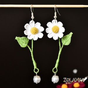 Op de afbeelding: Gehaakte witte madeliefjes oorbellen met groene bladeren en een witte parel hanger. De oorbellen hangen aan een houten stok.